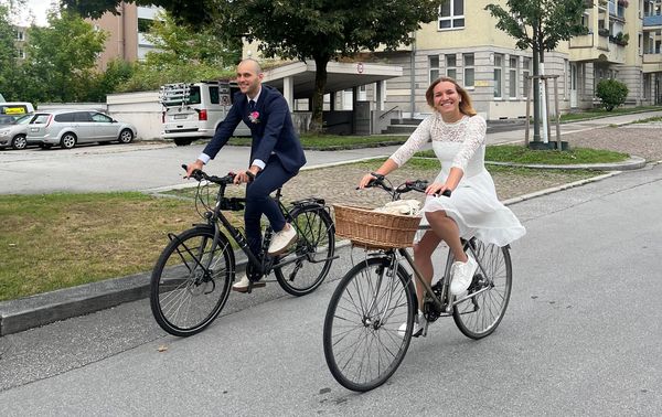 Serena and Philipp dressed in a white dress and a dark blue suit riding bicycles, smiling to the camera.